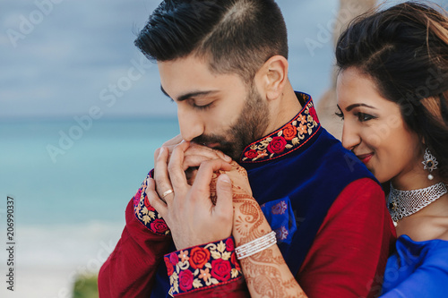 Wind blows around Hindu groom in blue sherwani and bride in lehenga posing in white house with gorgeous seaside view behind them photo