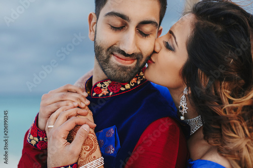 Wind blows around Hindu groom in blue sherwani and bride in lehenga posing in white house with gorgeous seaside view behind them photo