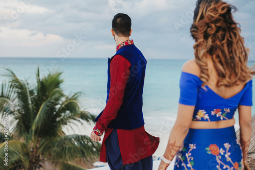 Wind blows around Hindu groom in blue sherwani and bride in lehenga posing in white house with gorgeous seaside view behind them photo