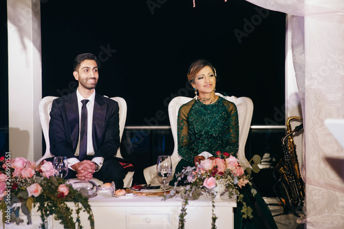 Indian groom in a classy black suit and beautiful bride in a green evening gown sit in the restaurant photo