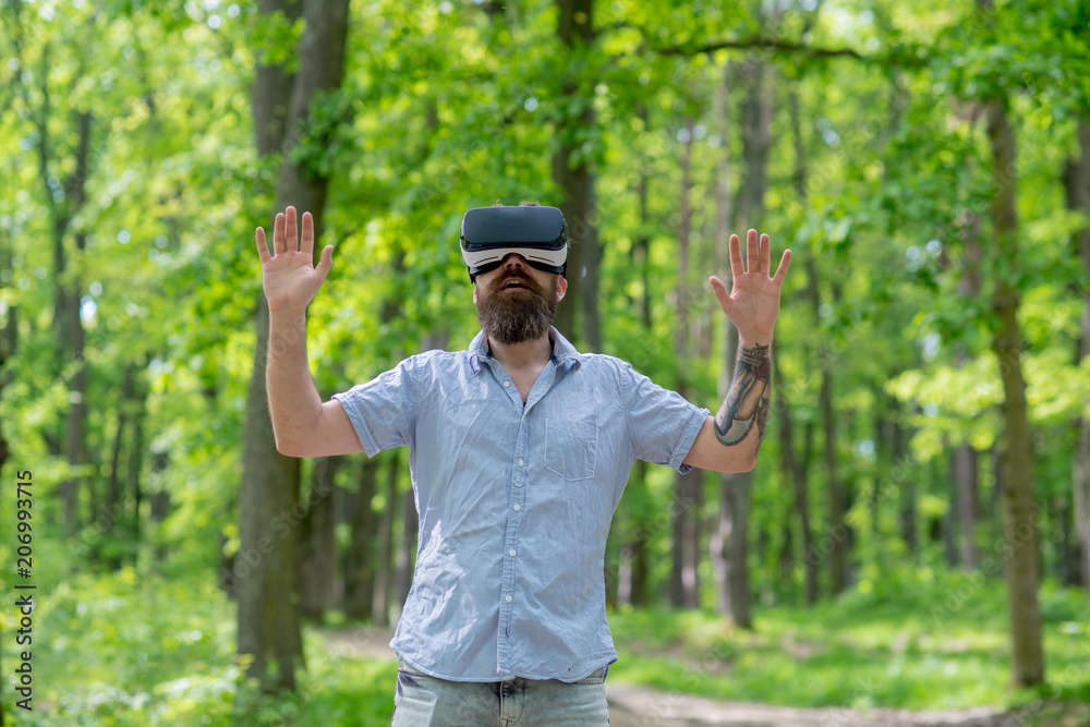 Bearded man wearing virtual reality glasses in forest. Smartphone using with VR goggles headset. Excited man with open mouth roaming in woods with modern gadget Bearded man wearing virtual reality glasses in forest. Smartphone using with VR goggles headset. Excited man with open mouth roaming in woods with modern gadget