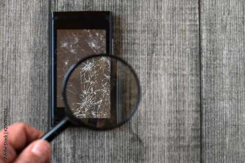 Broken Smart phone and magnifier in hand on gray wooden background.