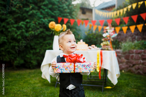 surprise for the birthday. the boy is holding a box with a gift in the yard on the background of a festive table with a cake. dressed in a black suit and a red butterfly