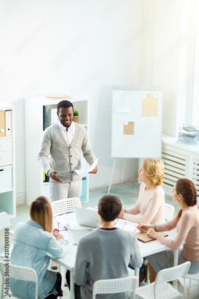 Smiling African American manager with sheet of paper in hands giving speech while participating in discussion of promising project, interior of modern boardroom on background Smiling African American manager with sheet of paper in hands giving speech while participating in discussion of promising project, interior of modern boardroom on background