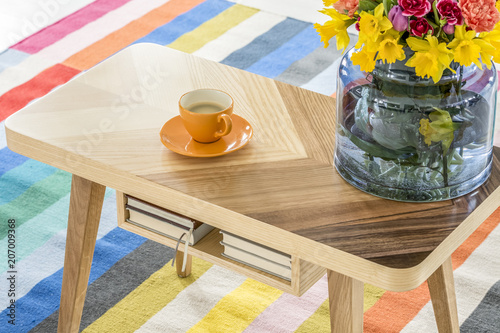 Close-up of a wooden table with a cup of coffee and a bouquet of spring flowers standing on a colorful striped rug