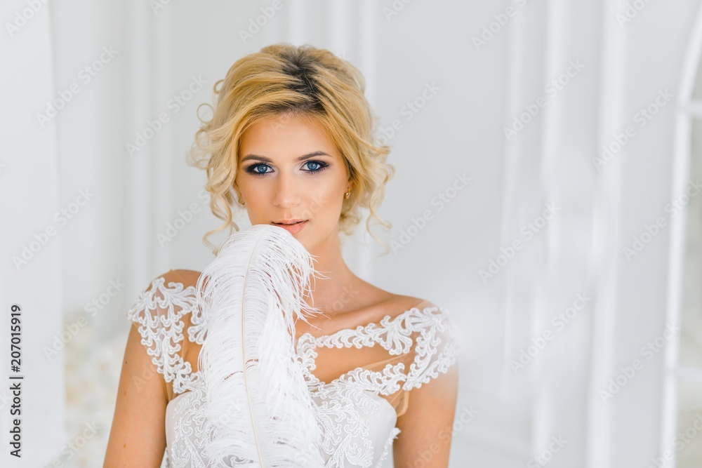 A bride with hairstyle and make up with white feahter. A portrait of beautiful girl with blond hair and blue eyes in wedding dress in studio A bride with hairstyle and make up with white feahter. A portrait of beautiful girl with blond hair and blue eyes in wedding dress in studio