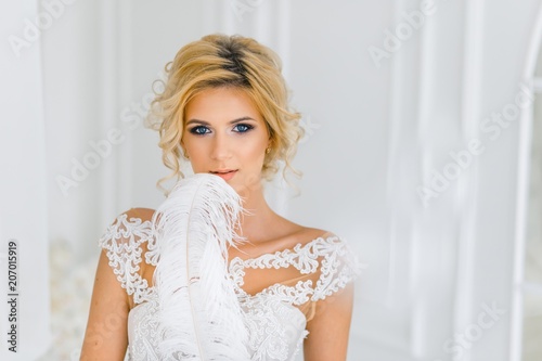 A bride with hairstyle and make up with white feahter. A portrait of beautiful girl with blond hair and blue eyes in wedding dress in studio