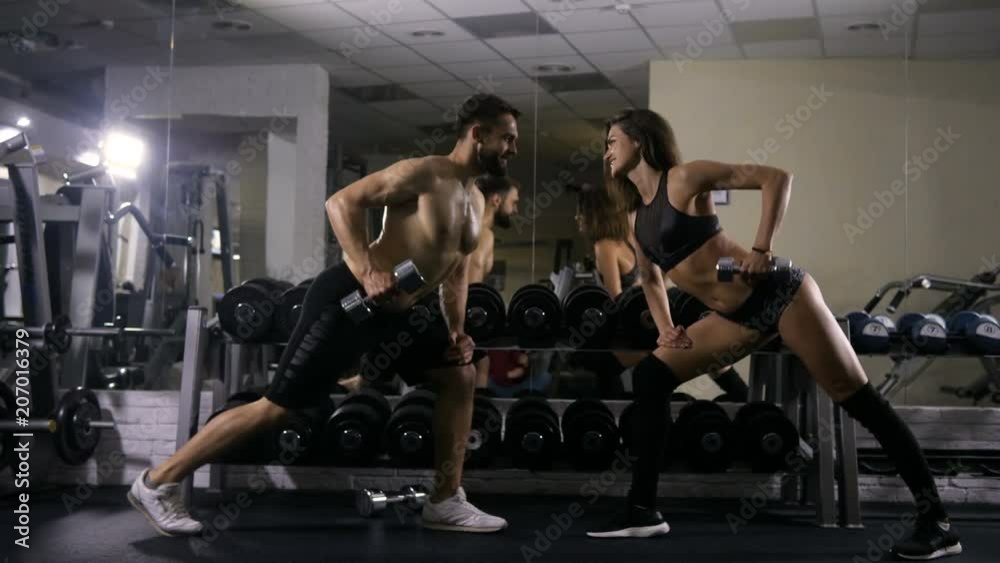The man and the woman of the sports body are engaged in the gym doing hand exercises with dumbbells standing one against the other, two trainings in the gym are practicing the exercises on the hands The man and the woman of the sports body are engaged in the gym doing hand exercises with dumbbells standing one against the other, two trainings in the gym are practicing the exercises on the hands