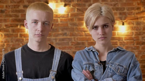 Young serious couple of hipsters is watching at camera, girl is crossinf her arms, brick background photo