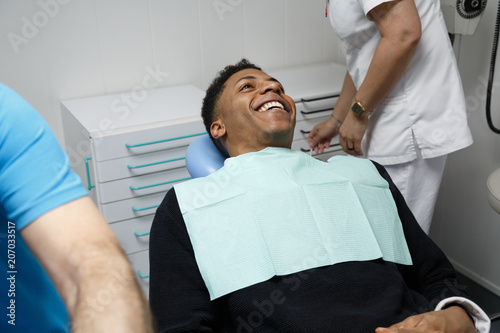Laughing African-American man sitting in chair of dentist in clinic and preparing for procedure with nurse.  