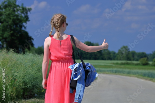 Młoda blondynka stoi (tyłem) na drodze z uniesionym kciukiem prawej ręki w znanym geście autostopowiczów, aby zatrzymać samochód, ma warkoczyki, różową letnią sukienkę bez rękawów, jeansową kurtkę 