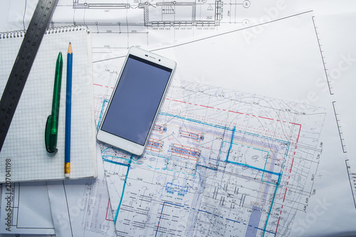 Green pen, blue pencil, white cellphone, metal ruler, notepad and some paper draws of a house (home) are laying down on white table. All staff is for a architecture negotiation.