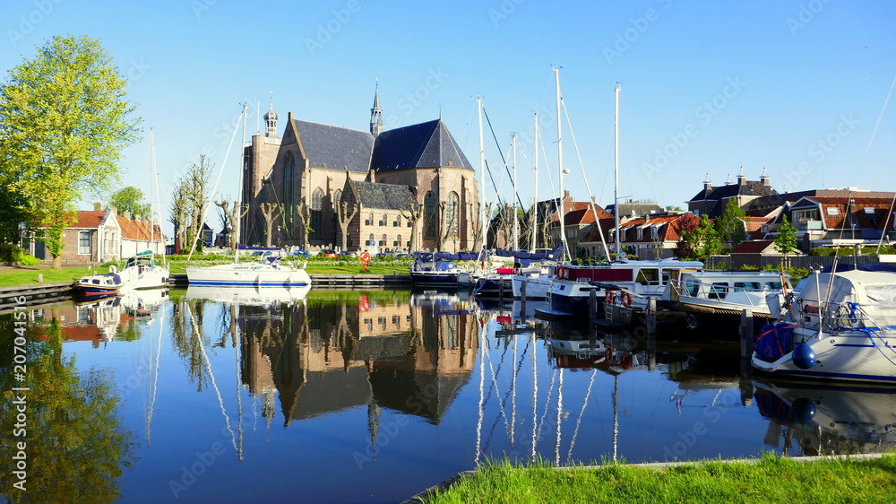 Am Hafen von Workum spiegeln sich die Kirche und die Schiffe im Morgenlicht im blauen Wasser.