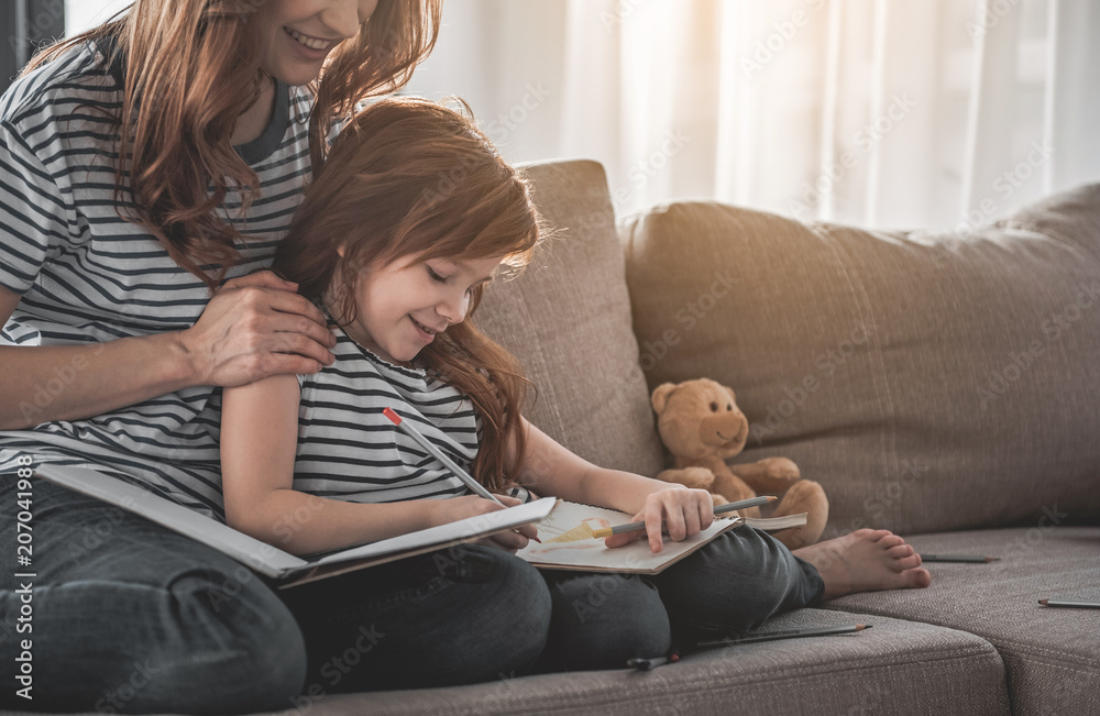 Family harmony. Portrait of smiling kid sitting on sofa with her mom holding her shoulders. Red-haired woman is watching her daughter with content. Pencils are scattered on sofa beside them