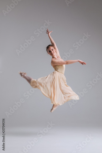 Enjoy moving your body with ease! Young cheerful attractive ballerina in jump showing split isolated on grey background.