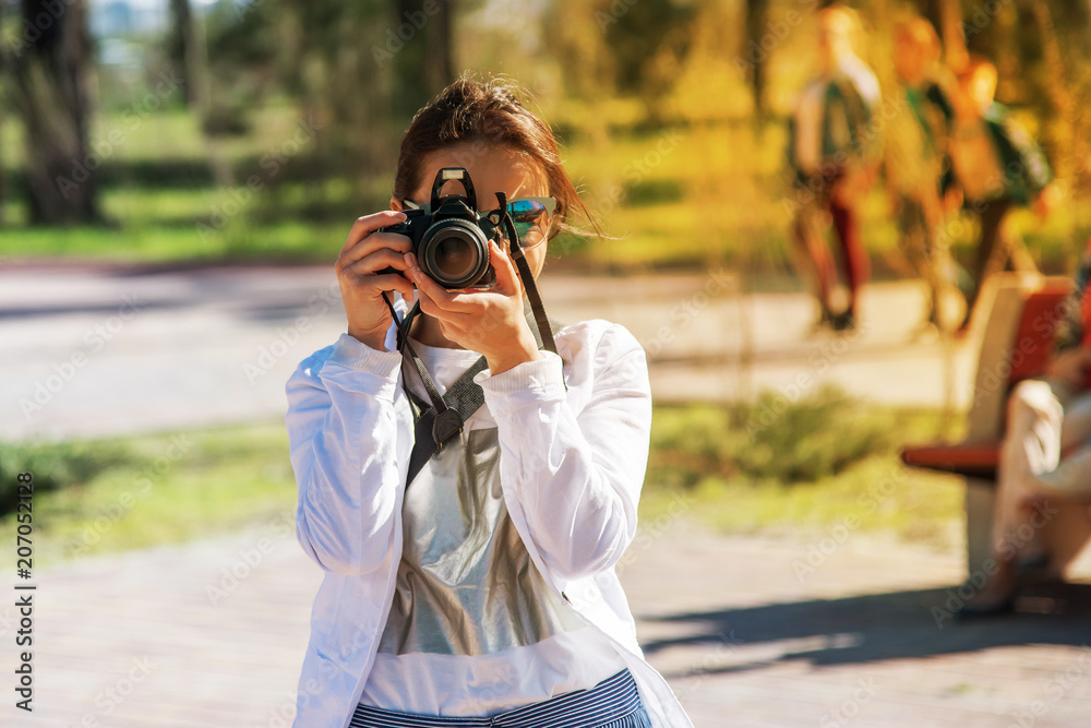 A girl in white clothes and blue glasses with a professional camera takes pictures on a bright sunny summer afternoon in the park