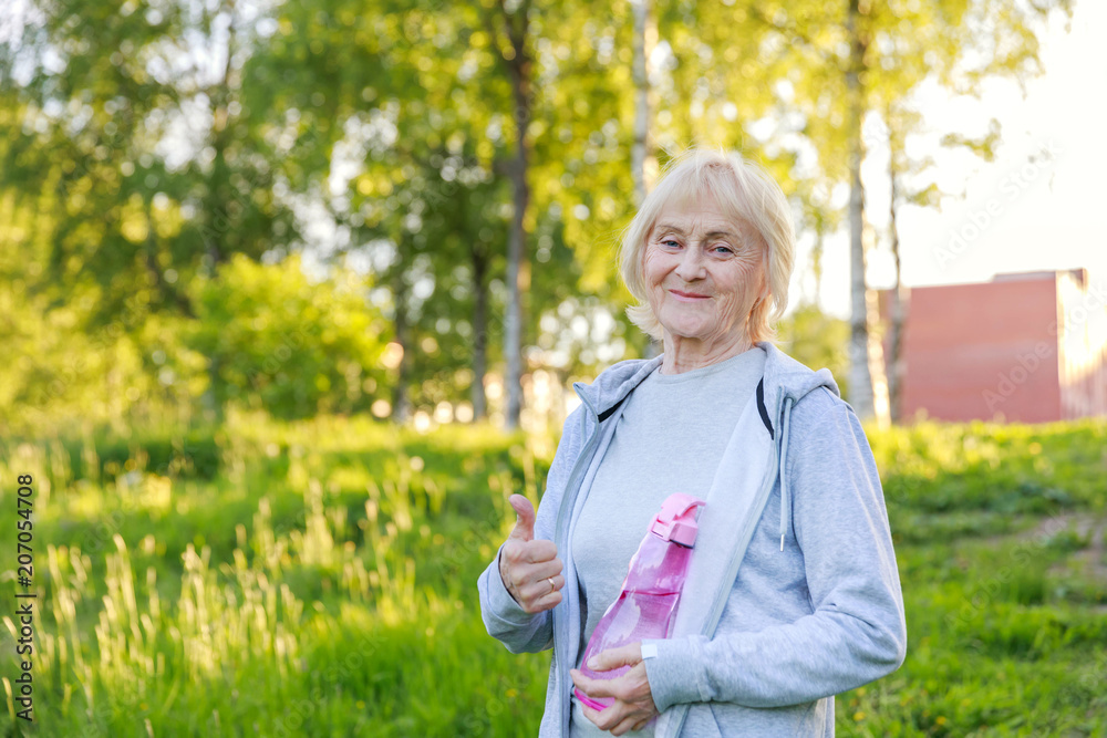 Happy elderly woman shows thumbs up in sportswear in a sunny park. Be active and energetic until old age, longevity, biohacking Happy elderly woman shows thumbs up in sportswear in a sunny park. Be active and energetic until old age, longevity, biohacking