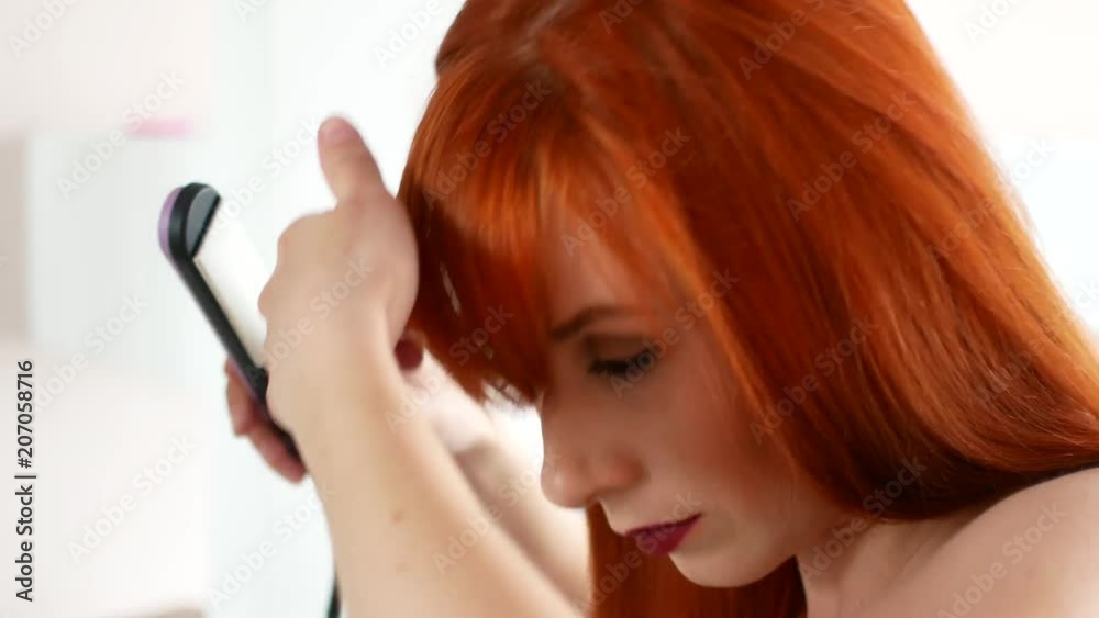 Red-haired woman balancing her hair with an iron before the mirror Red-haired woman balancing her hair with an iron before the mirror