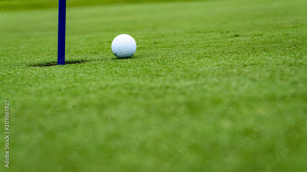 Side view of a golf ball on a sloped putting green, close to the hole with a blue pin
Side view of a golf ball on a sloped putting green, close to the hole with a blue pin