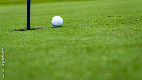 Side view of a golf ball on a sloped putting green, close to the hole with a blue pin 