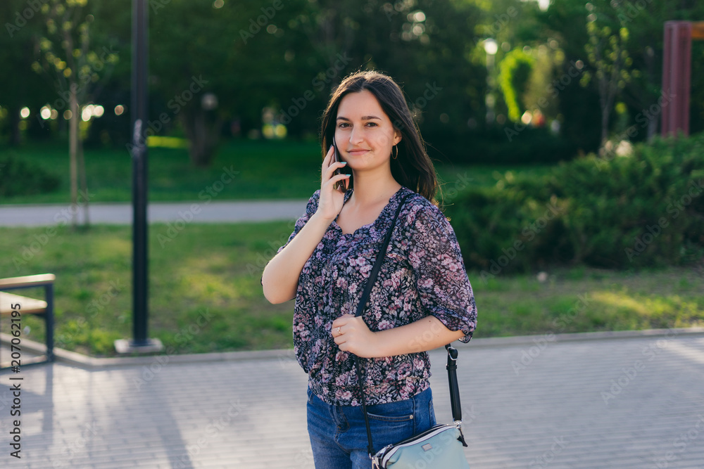 Young beautiful girl, talking on the phone, walking around the park. The girl walks the city on a warm sunny day Young beautiful girl, talking on the phone, walking around the park. The girl walks the city on a warm sunny day