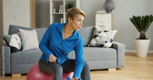Good looking middle aged woman in sporty clothes and with short fair hair in great shape sitting on the fitness ball and swinging her hands with dumbbells in the living room. Inside photo