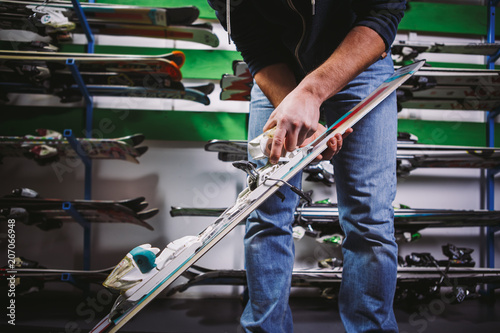 Theme of business sale and service of ski equipment. The hands of young Caucasian man have mountain skis, checks and adjusts the fastening for boot. On the wall of the stand with skis in the store photo