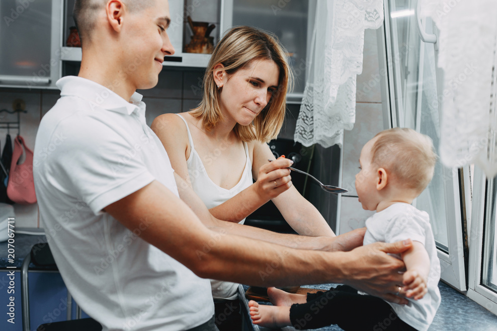 Fototapeta premium Mother feeding her baby daughter with spoon. Happy parents giving healthy food to her child at home kitchen. Childhood, parenthood, child care, development concept 