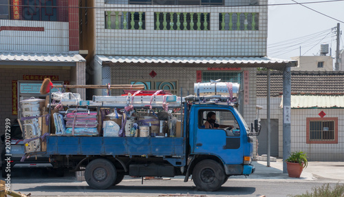 A itinerant truck is selling a lot of household items. This includes tables, mats, and ladders. The truck is blue. It is in front of a two st