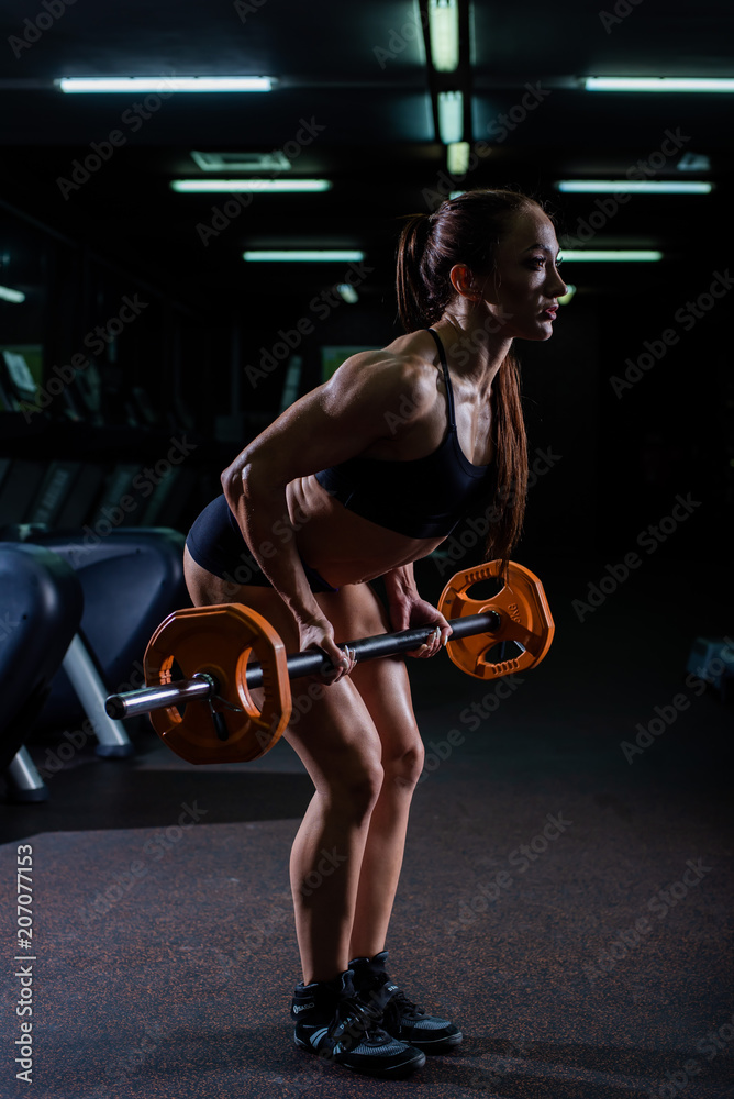 A strong, muscular, girl in black shorts, performs an exercise of deadlift, with a sports bar. Dark gym.