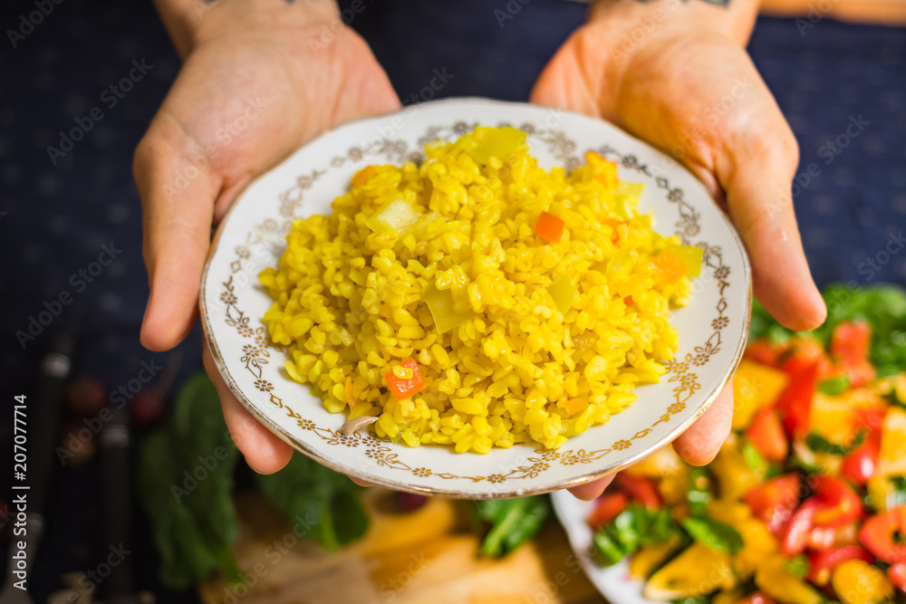 Woman hands holds cooked bulgur porridge pilaf or pilau served with raw vegetables salad. Yellow turmeric meal. Vegan vegetarian healthy food. Traditional Arabic asian food