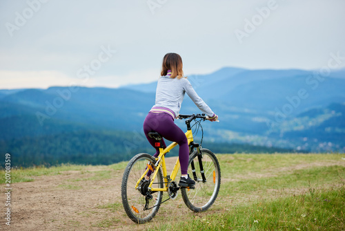 Back view of woman riding on yellow bicycle on a rural trail in the mountains on cloudy evening. Mountains, forests on the background. Outdoor sport activity, lifestyle concept