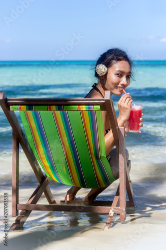 Ethnic girl with a white flower in her hair relaxed by the sea with red juice. Beautiful woman sitting in a deck chair on the sea beach. Tropical holidays on island.