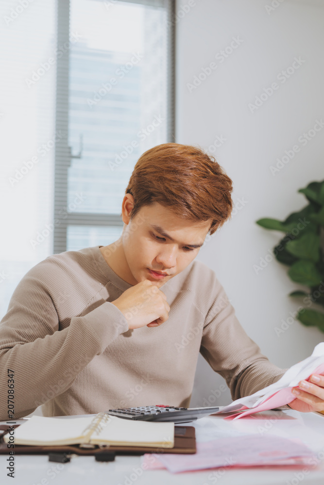 Unhappy asian man looking at bills sitting at table in living room Unhappy asian man looking at bills sitting at table in living room