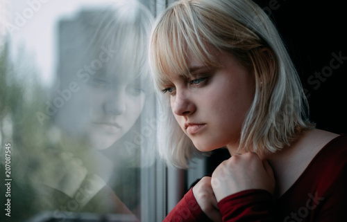 Portrait of depressed young blond woman near window at home. Sadness, nostlagic, depression.