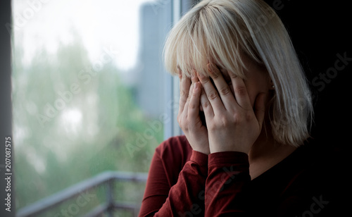 Portrait of depressed young blond woman near window at home. Sadness, nostlagic, depression. photo