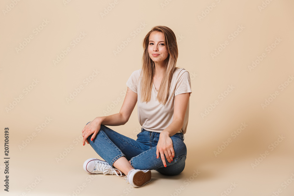Photo of pretty young woman wearing casual clothing smiing while sitting on floor with legs crossed, isolated over beige background Photo of pretty young woman wearing casual clothing smiing while sitting on floor with legs crossed, isolated over beige background