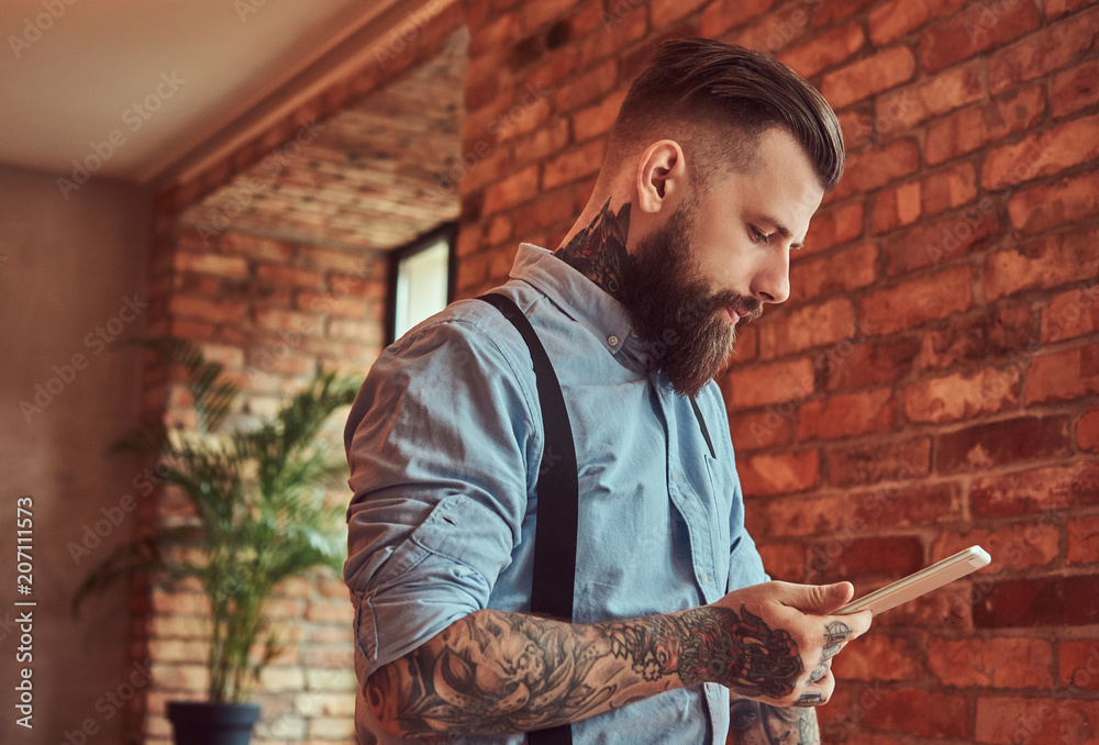 Fototapeta premium Old-fashioned tattooed hipster wearing a shirt and suspenders, in a sunglasses, using a tablet while standing near a desk in an office with loft interior.