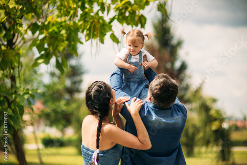 Happy joyful young family father, mother and little daughter having fun outdoors, playing together in summer park, countryside. Mom, Dad and kid laughing and hugging, enjoying nature outside © Aleksandr