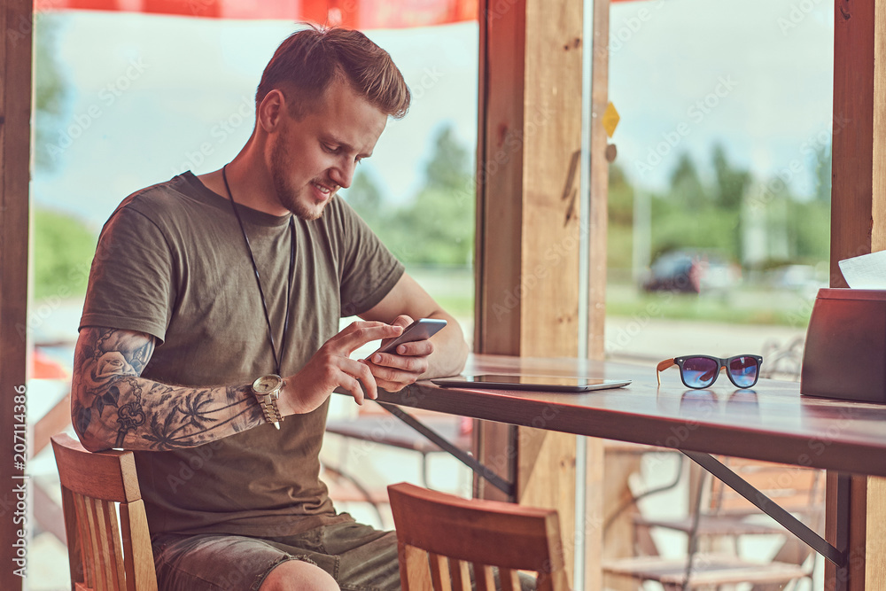 Handsome stylish hipster sits at a table in a roadside cafe, reading the message on the smartphone. Handsome stylish hipster sits at a table in a roadside cafe, reading the message on the smartphone.