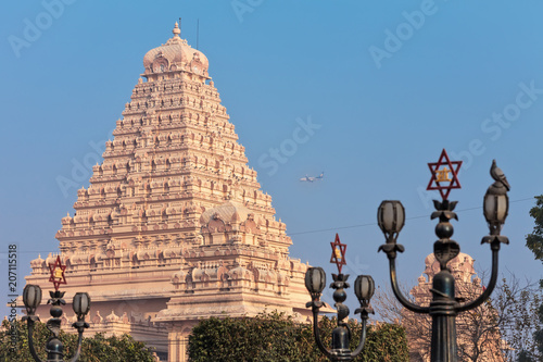 View of lamps with a traditional Hindu six-pointed star, and the main building of the Chattarpura temple complex near by which the passenger plane flies by.