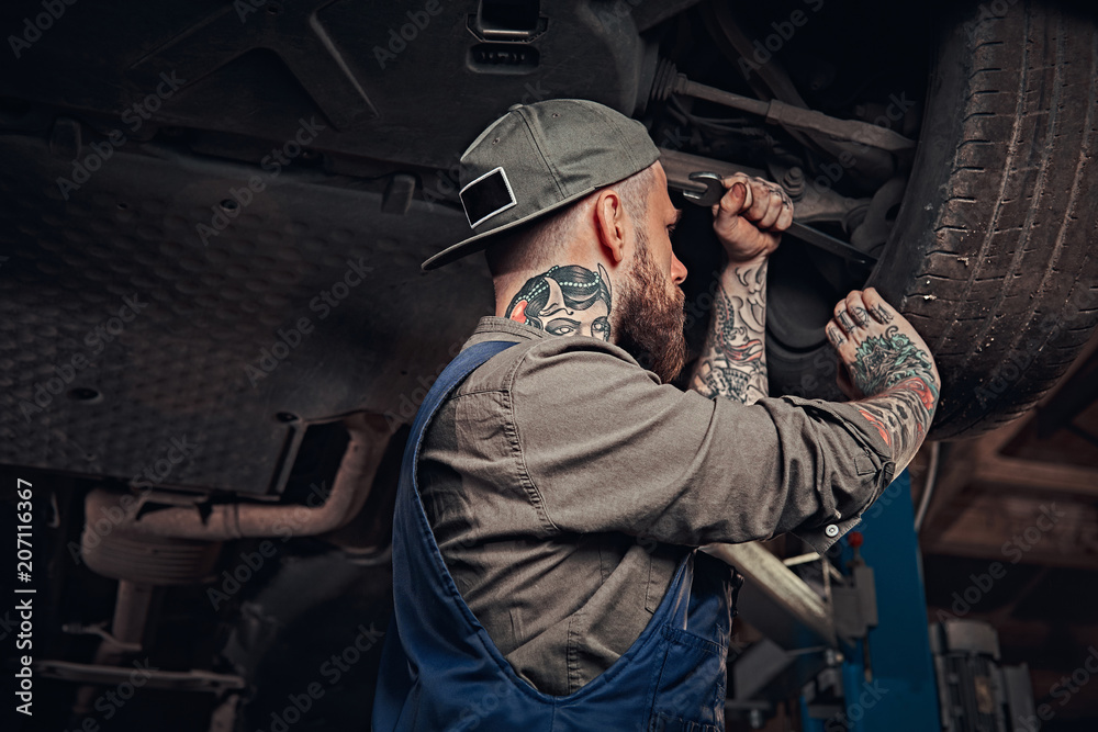 Bearded auto mechanic in a uniform repair the car's suspension with a wrench while standing under lifting car in repair garage. 