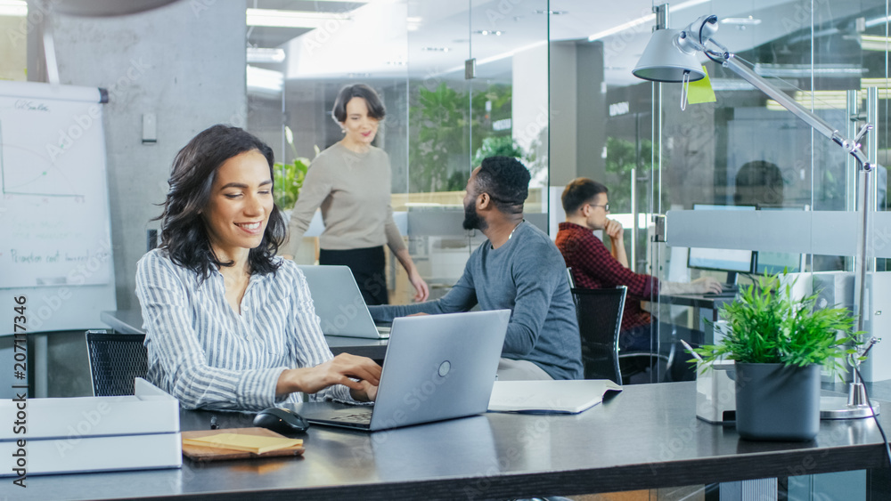 Busy International Office, Beautiful Hispanic Woman Working at Her Desk on a Laptop, in the Background Her Coworkers Have Work Related Conversations. Stylish Office with Bright Young People. Busy International Office, Beautiful Hispanic Woman Working at Her Desk on a Laptop, in the Background Her Coworkers Have Work Related Conversations. Stylish Office with Bright Young People.
