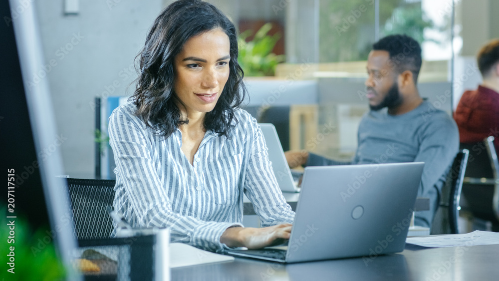 Busy International Office, Beautiful Hispanic Woman Working at Her Desk on a Laptop, in the Background Diverse Group of Creative Colleagues Working. Busy International Office, Beautiful Hispanic Woman Working at Her Desk on a Laptop, in the Background Diverse Group of Creative Colleagues Working.