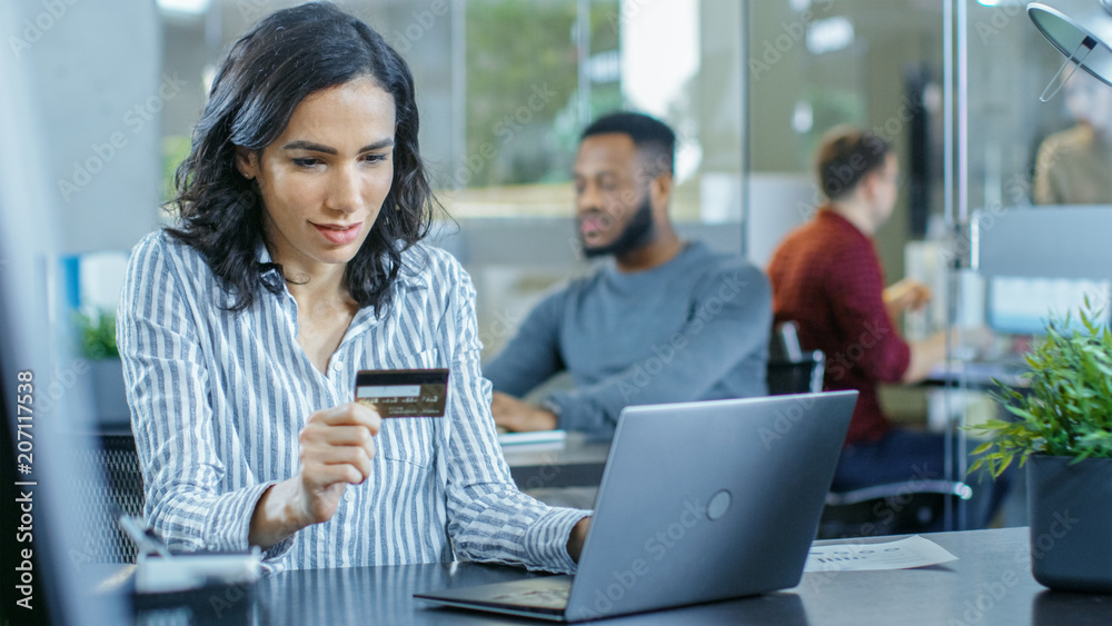 Fototapeta premium Beautiful Female Office Worker Makes Internet Purchase from Her Laptop Using Credit Card. In the Background Creative Young People Working.