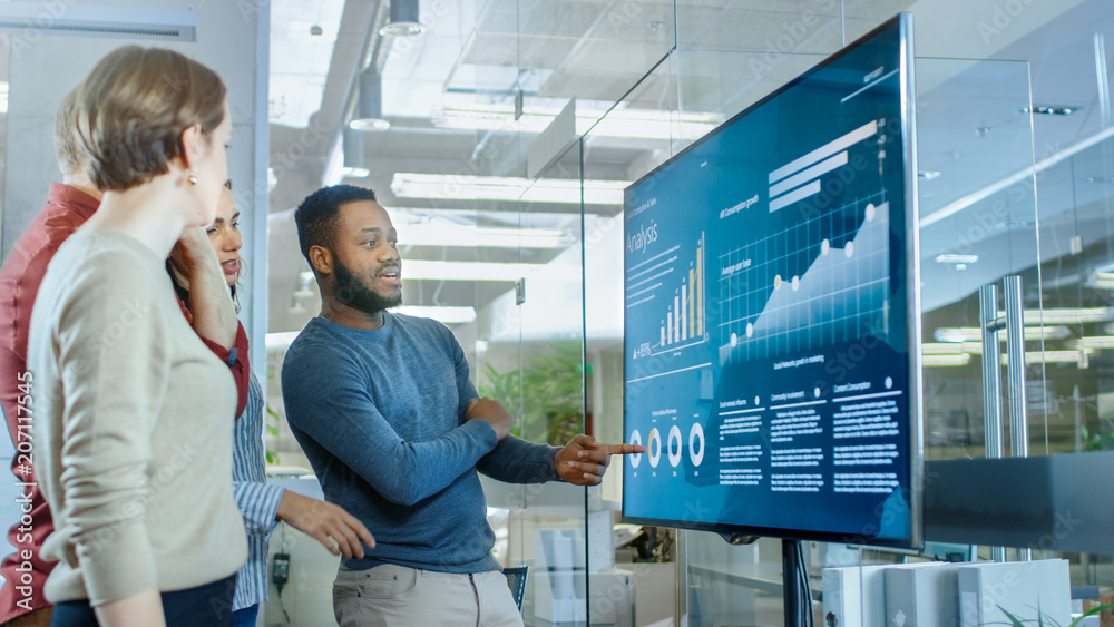 Diverse Team of Young Professionals in Conference Room Have Discussion about Statistics and Graphs Shown on a Presentation TV. Diverse Team of Young Professionals in Conference Room Have Discussion about Statistics and Graphs Shown on a Presentation TV.