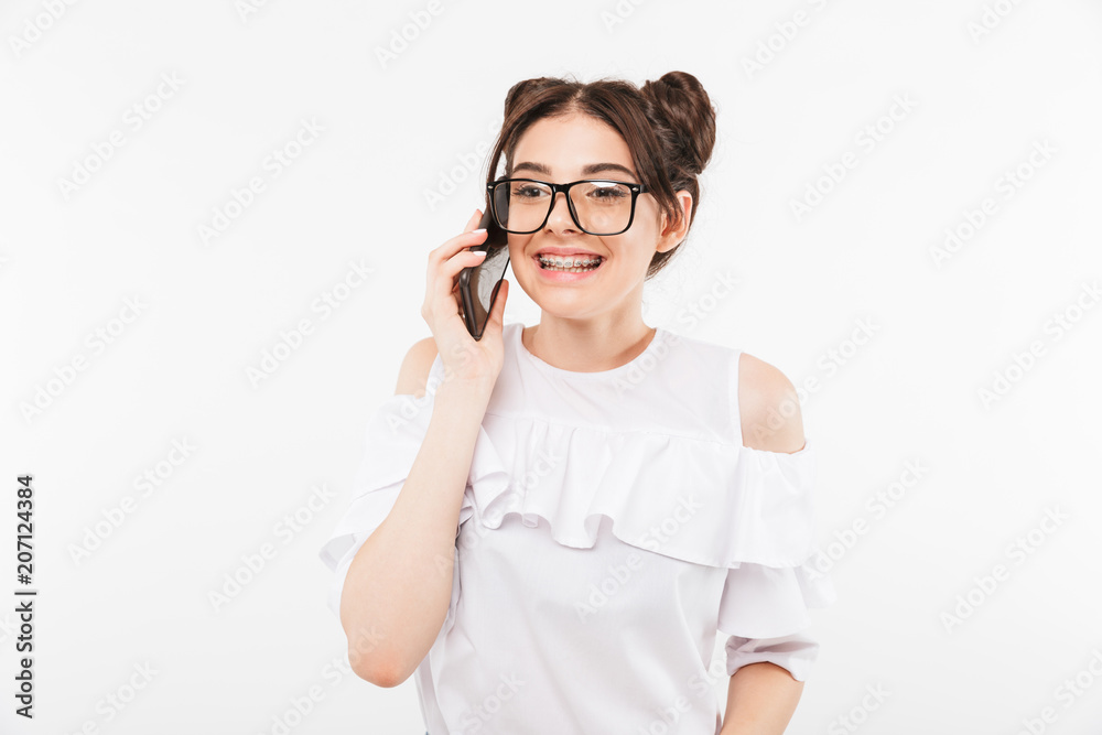 Image of happy teenage girl wearing two buns hairstyle and eyeglasses speaking on mobile phone and smiling with dental braces on teeth, isolated over white wall Image of happy teenage girl wearing two buns hairstyle and eyeglasses speaking on mobile phone and smiling with dental braces on teeth, isolated over white wall