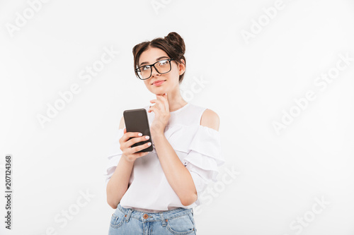 Photo of dreaming woman with double buns hairstyle looking upward and touching chin while holding smartphone, isolated over white background