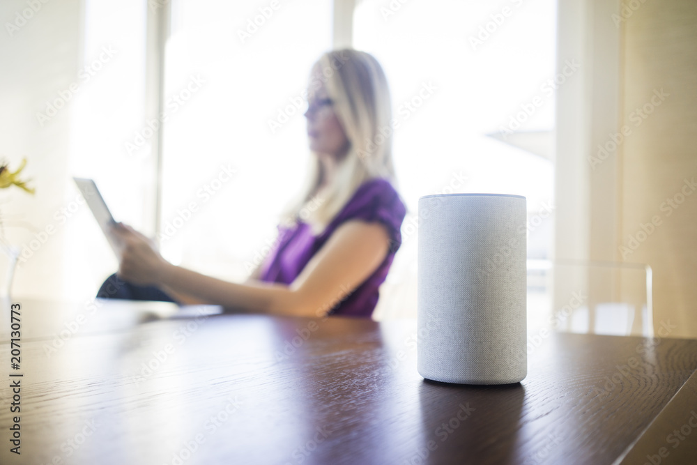 Smart Home loudspeaker on table with woman using tablet in the background Smart Home loudspeaker on table with woman using tablet in the background