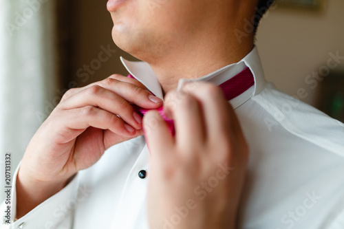 Elegant young fashion man dressing up for wedding celebration. Beautiful groom in a white shirt and a pink bow tie, preparing to wear a suit, preparing for the event. Man straightens his tie.